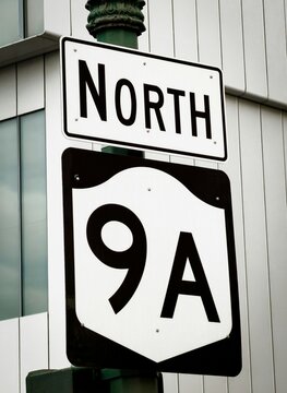Road sign with the word &ldquo;NORTH&rdquo; above a highway shield marked &ldquo;9A&rdquo; mounted on a pole. Urban exterior scene with clean typography, metal sign plates, and building facade in the background.