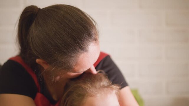 Mother kissing daughters head closeup, homemaker in apron shows tender affection at home, intimate soft focus shot, quiet protective embrace, warm living room atmosphere, soothing family connection