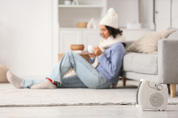 Obraz premium Electric fan heater on floor against woman drinking tea at home, closeup