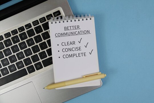 Notepad reading &ldquo;Better Communication&rdquo; with checklist items clear, concise and complete sits on a laptop keyboard beside a pen on blue background, conveying workplace communication and productivity.