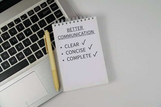 Notepad reading &ldquo;Better Communication&rdquo; with checklist items clear, concise and complete sits on a laptop keyboard beside a pen on white background, conveying workplace communication and productivity.