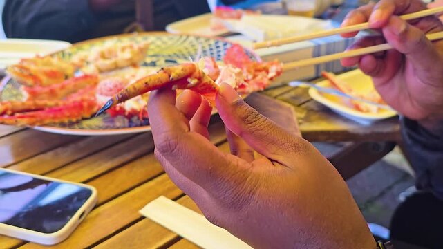 Hands using chopsticks to hold cracked king crab claw, enjoying fresh seafood from the market served on decorative plate on wooden table in Hakodate, Japan