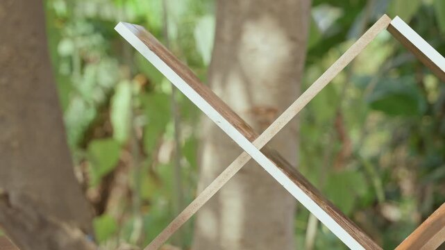 Crossed wooden slats near tree, carpentry trellis closeup showing peeling white paint on edges, diagonal pattern and textured grain with blurred foliage and sunlit bark, nostalgic outdoor mood