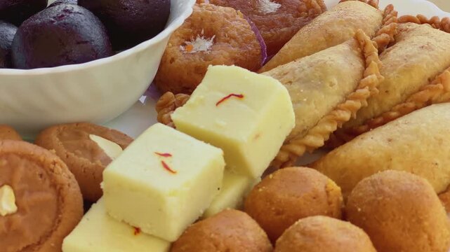 Turntable shot of an assorted selection of traditional Indian sweets including gulab jamun, peda, barfi, and gujiya, beautifully arranged on a plate with rich textures and warm tones