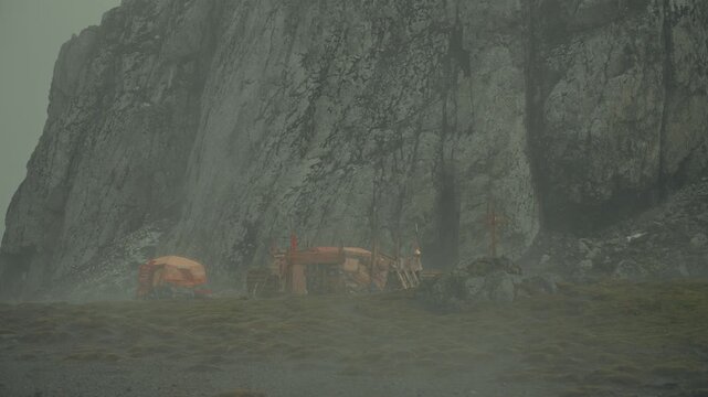 Makeshift Russian research base with orange shelters huddles at base of towering sheer cliff at Point Wild, Elephant Island, barely visible through swirling geothermal mist and harsh weather