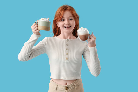 Teenage redhead girl with cups of sweet cocoa on blue background