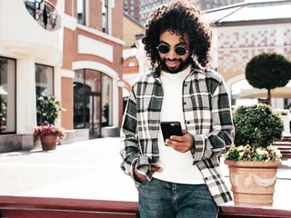 Fototapeten Friseursalon Handsome smiling hipster  model. Sexy unshaven Arabian man dressed in summer clothes. Fashion male with long curly hairstyle using smartphone apps, looking at cellphone screen. Holds phone  © halayalex