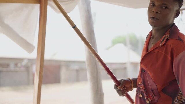 Young apprentice handling long wooden pole inside makeshift workshop under fabric canopy, steady hands measure and adjust timber for simple shelter construction, sunlit outdoor backdrop, focused