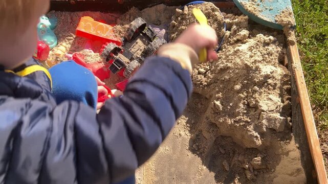 Over-the-shoulder view of little boy using yellow shovel to dig in wooden sandbox