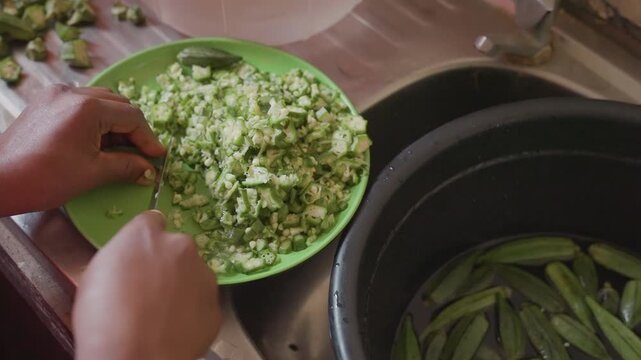 Closeup black hands transferring chopped okra into pot, communal kitchen energy, steady spoonless transfer, fresh pieces meet simmering vessel, warm family meal preparation scene