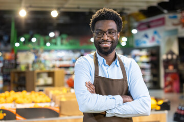 Fototapeta premium Young african american grocery store owner wearing an apron, arms crossed and smiling confidently at the camera while managing his small business among fresh produce