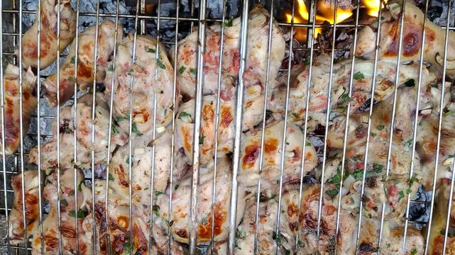 Pieces of meat, clamped in a metal grate and grilled over coals. The meat is covered in a marinade with herbs and spices. Gray, hot coals are visible from below.