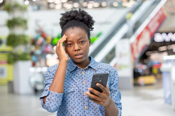 Young woman standing in a shopping mall, holding a mobile phone and touching her head, expressing feelings of headache, stress, and frustration from bad news or a difficult situation