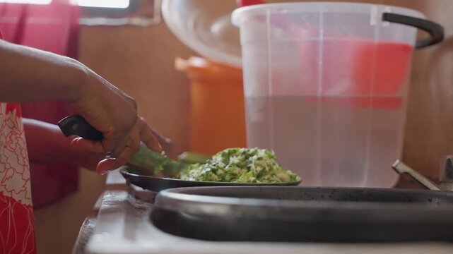 Black woman transferring chopped okra into large plastic container near sink, hands scooping with small spatula, pile of greens on plate, soft window light, preparation for storage or group meal,