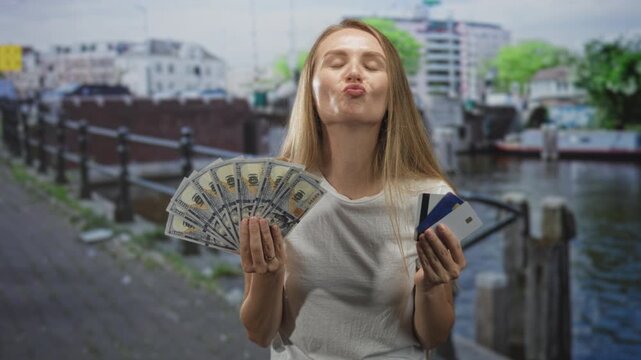 Woman holding fan of us dollars and creditcard in hands on street, puckers lips while presenting cash and cards; playful confidence.