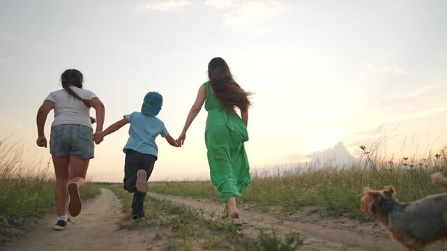 Three children run holding hands on dirt path at sunset with dog following. Boy and girls play outdoors in countryside. Kids enjoy freedom with terrier. Children run together. Sunset play with dog.