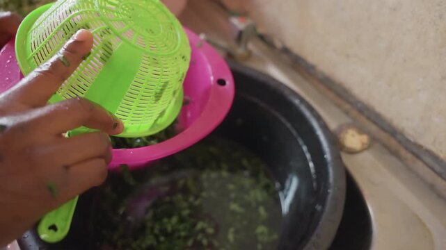 Black home cook rinsing chopped greens in green colander over sink, closeup of wet hands, herbs falling through sieve into basin, gentle motion, kitchen countertop and pink sieve visible, domestic