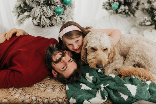 Young girl sits on floor with dad and dog in front of flocked trees