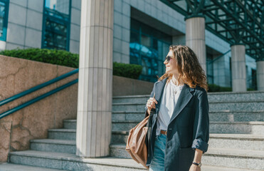 Fototapeta premium Stylish young businesswoman in jacket and sunglasses leaving office building in bright sunlight