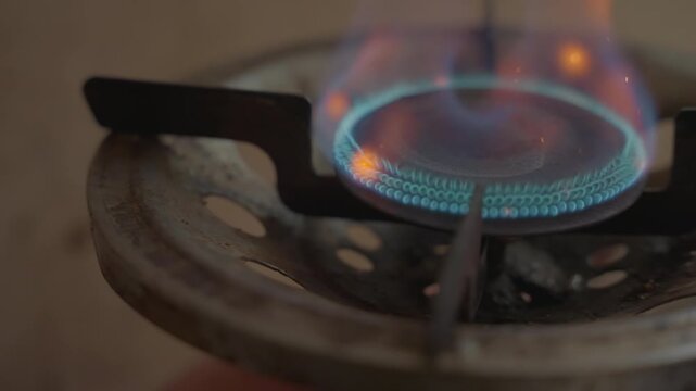 Blue gas flame dancing on burner, turquoise ring glows, rusted grate in foreground, soft orange flecks, steady pilot, ambient kitchen light, cinematic slow motion feel, textured metal, quiet domestic