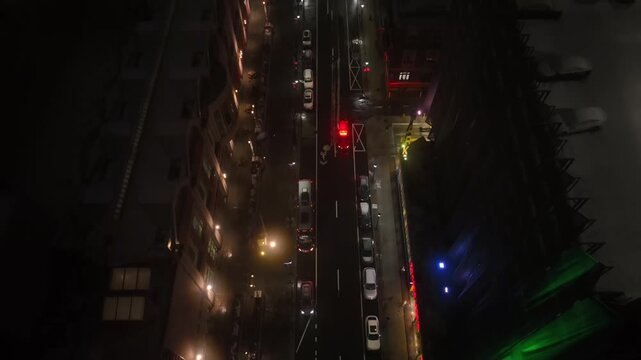 Aerial night tracking shot over snowy city street with ambulance lights flashing, traffic moving between buildings and wet roads reflecting urban glow in cold winter atmosphere. Tilt down.
