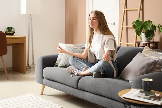 Young pretty woman meditating on sofa in stylish living room