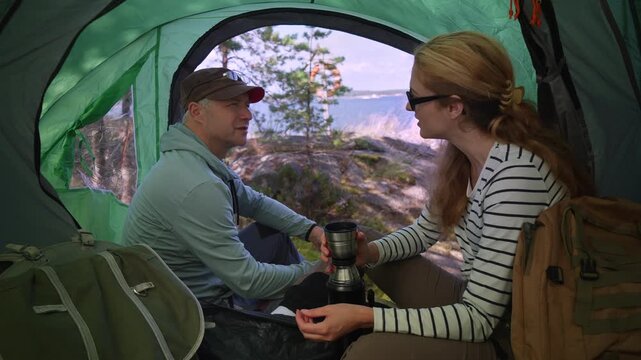 Adult couple talking and drinking tea inside a tent with a scenic coastal view. Authentic camping lifestyle, connection, and slow travel adventure during summer vacation.