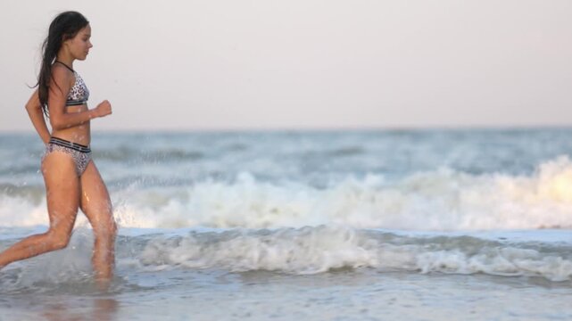 A girl with wet hair in a leopard swimsuit runs along the sea with waves peering into the horizon