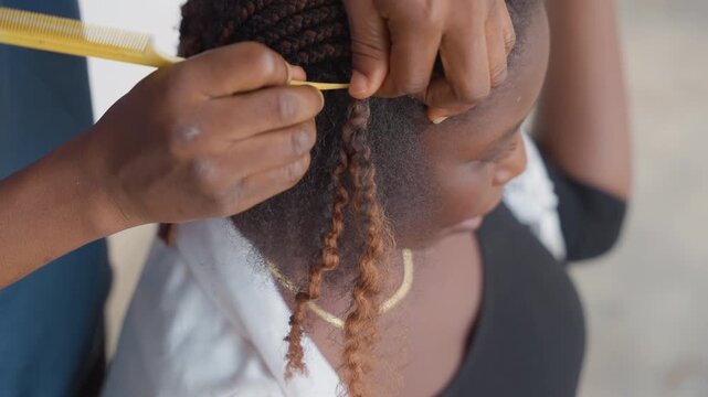 Medium shot black hairdresser braiding child hair outdoors, comb and extension used, gold necklace visible, focused hands creating neat parts, calm atmosphere, skillful protective styling lesson