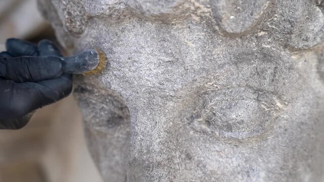 Restorer in black glove and lab coat using brass wire brush to clean patina from weathered stone sculpture. Professional conservation of historic landmark facade.