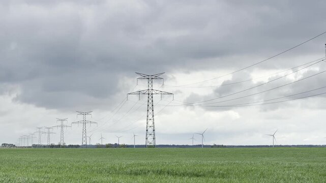 Perspective view where a row of transmission poles guides the eye across a green wheat field toward distant wind turbines, under a soft clouded sky shaping the rural energy landscape