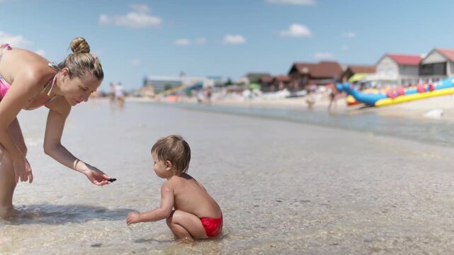 The kid collects shells and pebbles in the sea on a sandy bottom with his mother under the summer sun on vacation