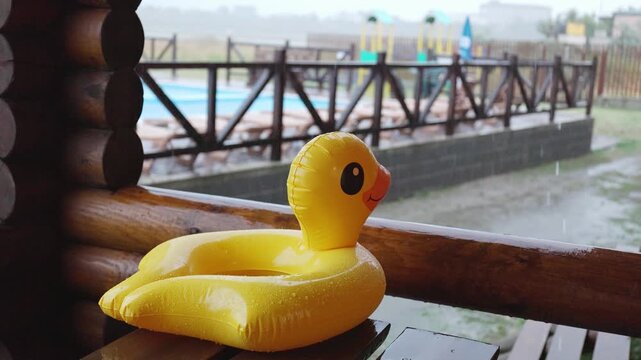 An inflatable duck lies on wooden table in gazebo at recreation center against the backdrop of pool covered with rain