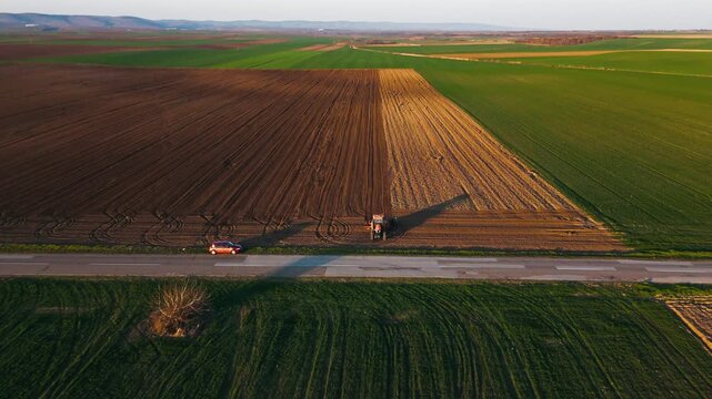 Aerial view of a tractor tilling soil, creating a stark contrast with green fields alongside a road with a car, Jazak, Vojvodina, Serbia.