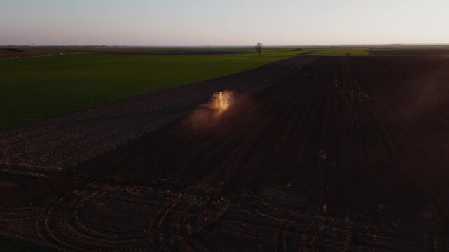 Aerial view of a tractor tilling the dark soil of a vast field, kicking up a cloud of dust against the setting sun, Jazak, Vojvodina, Serbia.