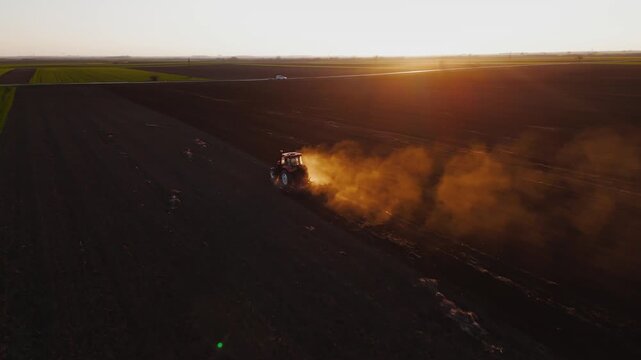 Aerial view of a tractor kicking up dust as it tills the soil in a vast field with vivid green patches, Jazak, Vojvodina, Serbia.