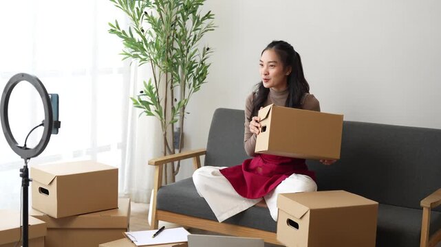 Young asian businesswoman using a smartphone on a tripod with a ring light to live stream while promoting her products, showing cardboard boxes from her home office, working as a vlogger