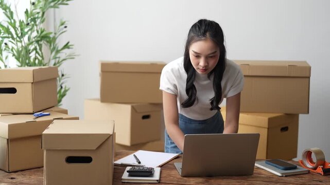 Young asian business woman using a laptop computer to check online orders and manage her e-commerce business from a home office surrounded by cardboard boxes ready for shipping