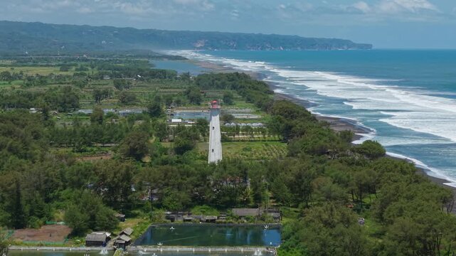 Aerial view of the Mercusuar Samas lighthouse, standing tall amidst lush greenery, adjacent to the dark sandy beach and ocean, Bantul Regency, Indonesia.
