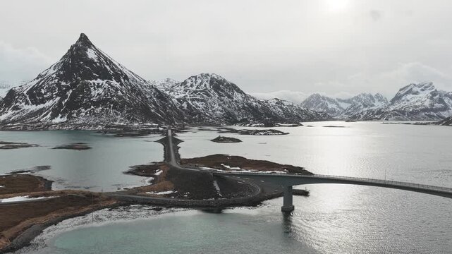 Aerial view of the Flakstadbruene bridges cutting through the waters surrounded by snow-capped mountains under a cloudy sky, Flakstadbruene, Nordland, Norway.