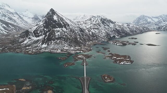 Aerial view of the bridge connecting islands amidst the turquoise waters contrasting with the snow-capped mountains, Flakstadbruene, Nordland, Norway.