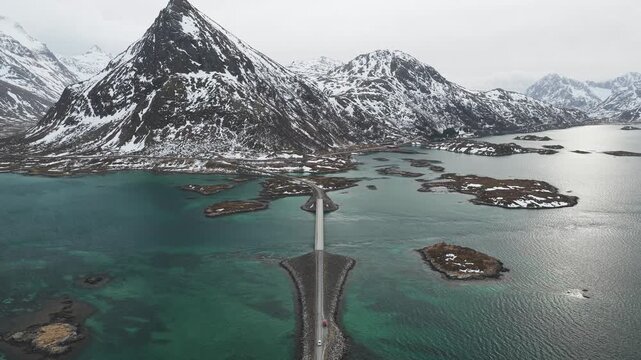 Aerial view of the Flakstadbruene bridge cutting through the turquoise water, connecting islands beneath snow-capped mountains, Flakstadbruene, Nordland, Norway.