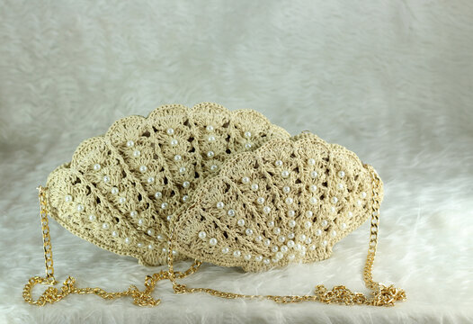 Beige crocheted shoulder bag in the shape of a seashell with a gold-colored handle sits on a white fluffy blanket. The bag features pearl bead details.