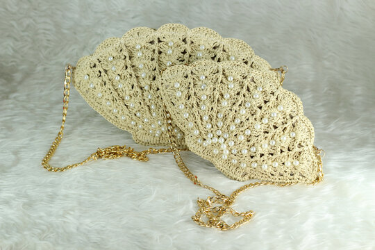 Beige crocheted shoulder bag in the shape of a seashell with a gold-colored handle sits on a white fluffy blanket. The bag features pearl bead details.