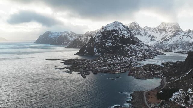 Aerial view of majestic snow-capped mountains overlooking the quaint village nestled along the rugged coastline, Tind, Nordland, Norway.