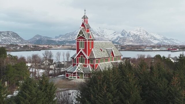 Aerial view of the striking red Buksnes Church stands out against the backdrop of snow-capped mountains and a tranquil lake, Buksnes Kirke, Nordland, Norway.