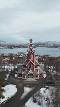 Aerial view of the red Buksnes Church with its detailed architecture standing out against the snowy landscape and grey sky, Buksnes Kirke, Nordland, Norway.