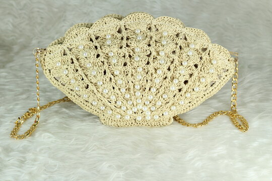 Beige crocheted shoulder bag in the shape of a seashell with a gold-colored handle sits on a white fluffy blanket. The bag features pearl bead details.