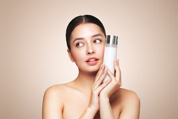 Smiling woman holds bottle with cosmetic milk. Photo of attractive woman with perfect makeup on beige background. Beauty concept