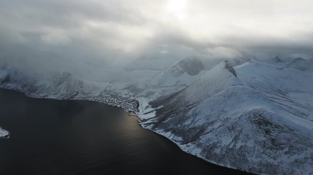 Aerial view of Segla mountain peak covered in snow, meeting the dark waters under a cloudy sky, creating a stark, beautiful contrast, Segla, Troms, Norway.